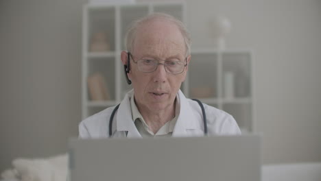 Un-Anciano-Profesor-De-Medicina-Está-Usando-Una-Computadora-Portátil-Para-Consultar-En-Línea-A-Pacientes-Y-Estudiantes-Internos-Desde-Su-Oficina
